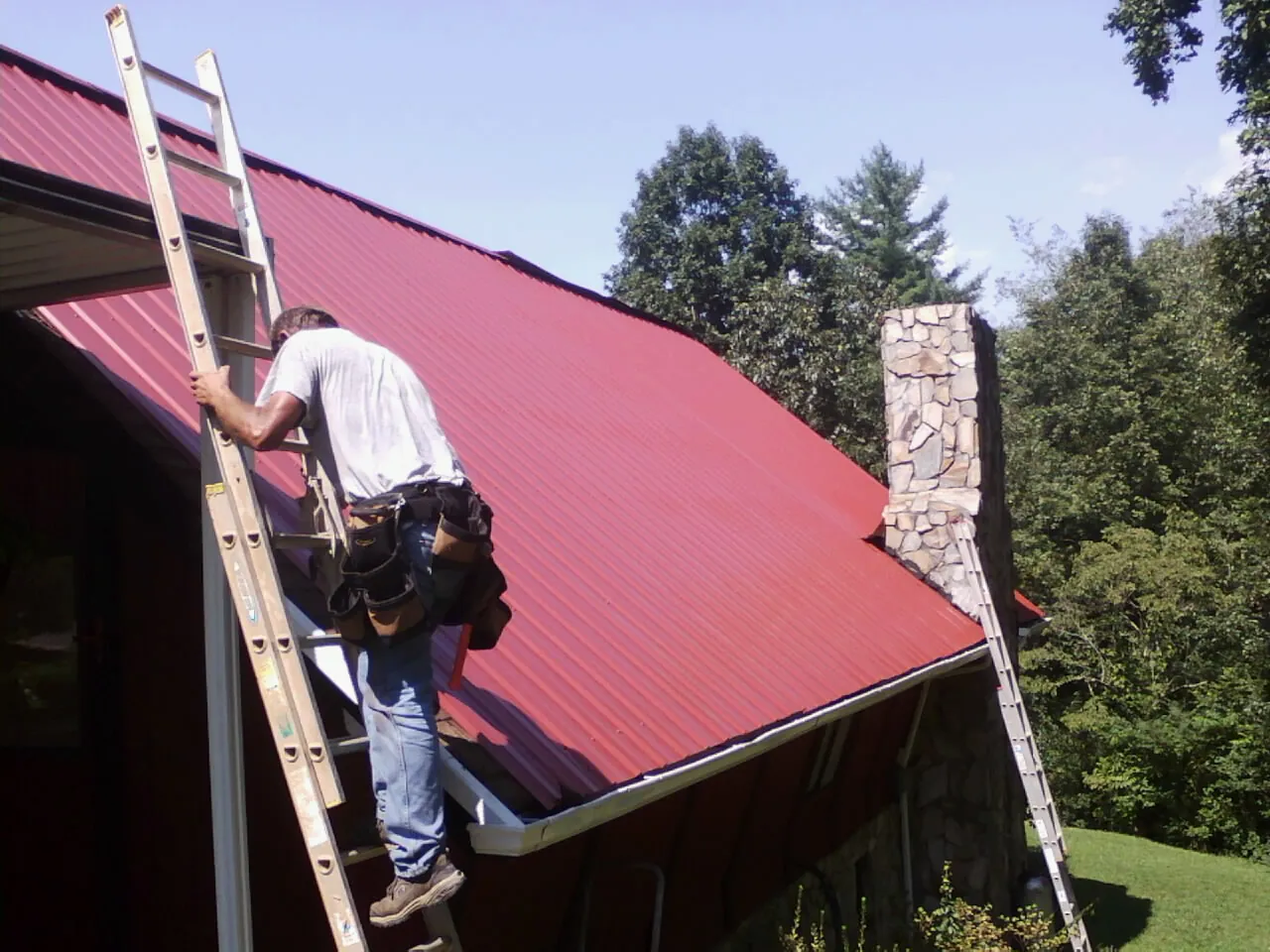 Roofer on a ladder beside a red metal roof and stone chimney — Soffit & Fascia Repair by Roofing Brothers in Croton